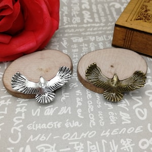 Op de afbeelding: Twee metalen vogelbedels, een zilveren en een bronzen, worden getoond op houten bases. De vogels bevinden zich in een vliegende positie met gespreide vleugels.
