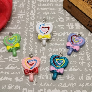 May include: Four colorful heart-shaped lollipop charms with bows. The lollipops are green, white, purple, and blue. Each charm has a silver loop for attaching to a necklace or bracelet.
