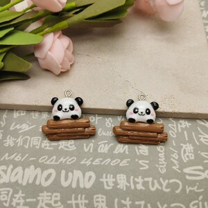 May include: Two panda bear charms. Each panda has a black and white face with a smiling expression and sits on a stack of brown logs. The charms have a silver-colored loop at the top for attaching to jewelry or other crafts.