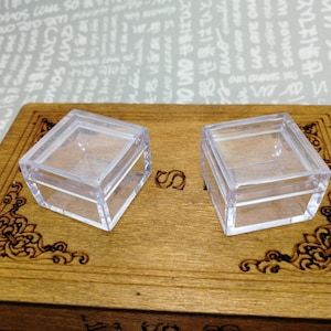 Puede incluir: Dos cajas cuadradas de plástico transparente con tapas sobre una superficie de madera con un diseño ornamentado.