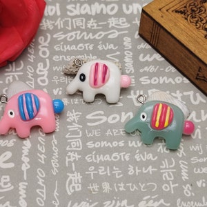 Pode incluir: Três pequenos encantos de elefante feitos de argila polimérica. Os elefantes são rosa, branco e verde com listras e detalhes coloridos. Cada elefante tem um pequeno laço para prender a um colar, pulseira ou outra joia.