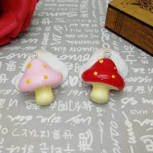 May include: Two mushroom-shaped charms. One is pink with yellow spots, the other is red with yellow spots. Both have a light yellow stem and a silver loop for attaching to jewellery or other crafts. The charms are on a patterned surface.
