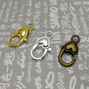 Könnte beinhalten: Drei herzförmige Karabinerverschlüsse in Gold-, Silber- und Bronze-Finish. Die Verschlüsse sind für die Schmuckherstellung und das Basteln konzipiert.