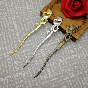 Puede incluir: Tres palitos decorativos para el pelo con diseños ornamentados. Los palitos son de color dorado, plateado y bronce. Los diseños presentan una cabeza de animal estilizada en la parte superior.