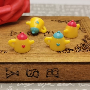 Puede incluir: Cuatro teteras amarillas en miniatura con detalles azules y rojos. Cada tetera tiene un pequeño diseño de corazón en la parte delantera. Las teteras están sobre una superficie de madera.