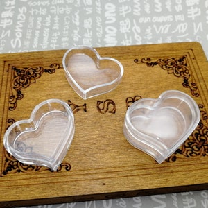 Puede incluir: Tres recipientes de plástico transparente en forma de corazón sobre una superficie de madera con un diseño decorativo. Los recipientes están apilados uno encima del otro.