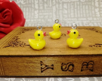 May include: Three yellow rubber duck charms with red beaks and black eyes. The charms are on a wooden surface with a decorative pattern.
