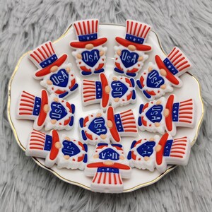 May include: A collection of 12 red, white, and blue gnome-shaped silicone charms with the word "USA" on each charm. The charms are on a white plate with a gold rim.