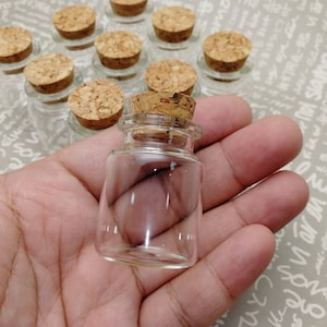 May include: A close-up of a small, clear glass bottle with a cork stopper. The bottle is sitting in the palm of a hand.