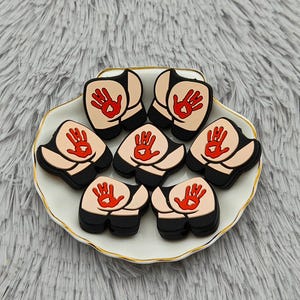 May include: Seven novelty shoe charms shaped like buttocks with a red handprint on each. The charms are black, tan, and red, arranged on a white plate with a gold trim. The plate sits on a fluffy, gray surface.