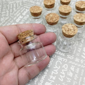 May include: A hand holding a small, clear glass bottle with a cork stopper. The bottle is empty and has a round shape. There are several other similar bottles in the background.