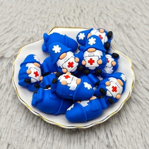 May include: A collection of blue gnome figurines in a white and gold-rimmed dish. Each gnome wears a blue hat with a white star, a white coat with a red cross, and a white beard. The gnomes are clustered together.