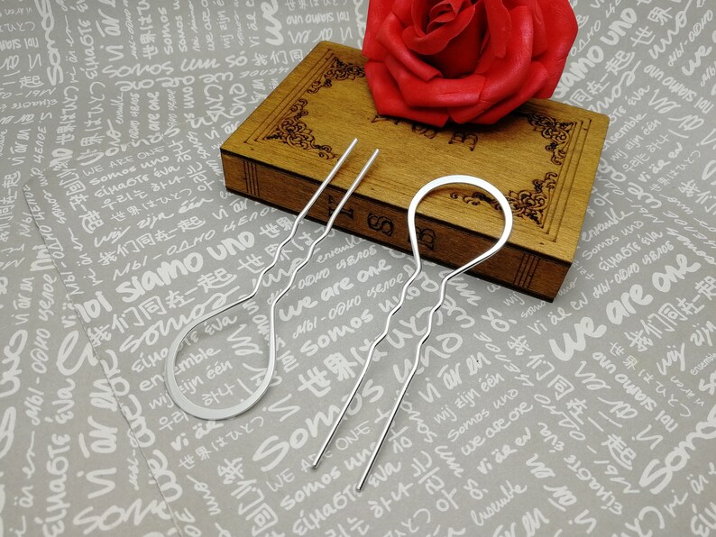 Puede incluir: Tres pinzas para el pelo plateadas con dise&ntilde;os ondulados. Las pinzas est&aacute;n sobre una superficie de madera con una rosa roja en el fondo.