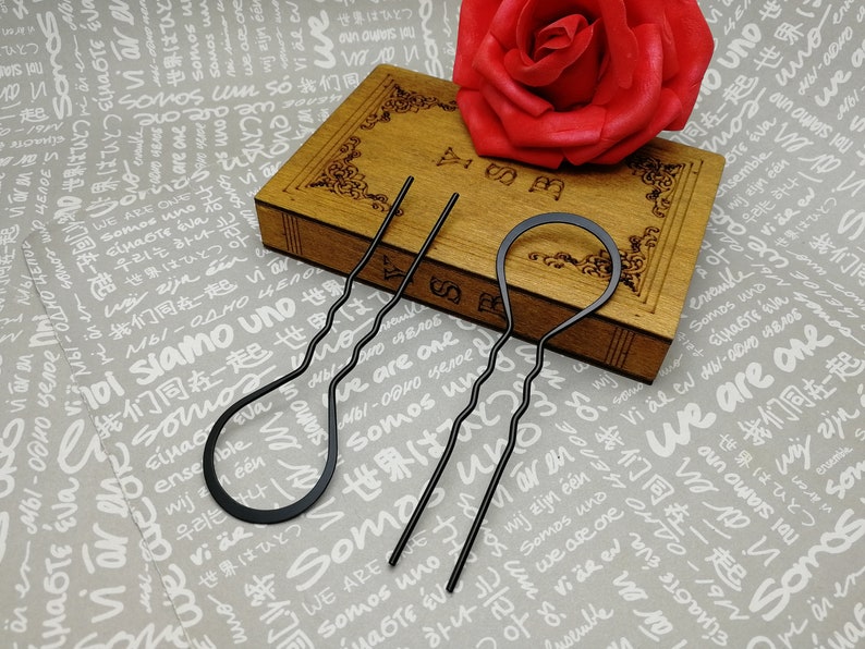 Puede incluir: Tres horquillas de metal negro. Dos de las horquillas tienen un dise&ntilde;o ondulado y un bucle en el extremo. La tercera horquilla es recta con un bucle en el extremo. Las horquillas est&aacute;n sobre una superficie de madera con una rosa roja en el fondo.