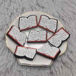 Puede incluir: Un conjunto de colgantes con forma de libro con las frases "we lose ourselves in books" y "we find ourselves there too". Los colgantes negros, blancos y rojos están colocados sobre un plato blanco con borde dorado.
