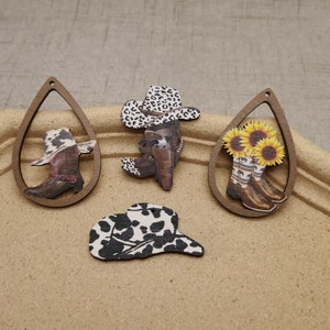 May include: Four wooden cutouts with different designs. One cutout is a cowboy hat with a black and white cow print. Another cutout is a pair of cowboy boots with sunflowers. The third cutout is a cowboy hat with a black and white cow print. The fourth cutout is a pair of cowboy boots with a black and white cow print.