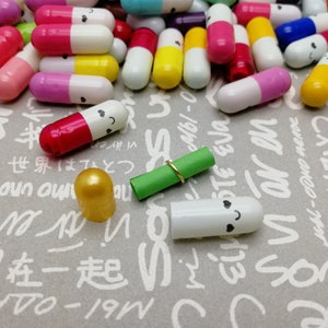 以下が含まれることがあります： 笑顔が印刷されたカラフルなカプセルのコレクション。カプセルは白い文字が書かれた灰色の表面に散らばっています。表面には金色のリングと金色のキャップが付いた緑色の巻かれた紙があります。