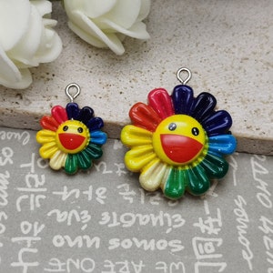 Puede incluir: Dos encantos de flores sonrientes de colores con pétalos de arcoíris y una cara amarilla. El encanto más grande tiene un diámetro de aproximadamente 5 cm y el encanto más pequeño tiene un diámetro de aproximadamente 2,5 cm.