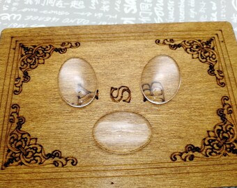 Pode incluir: Uma placa de madeira com três formas ovais de vidro transparente, duas na parte superior e uma na parte inferior. A placa tem um design decorativo ao redor das bordas.