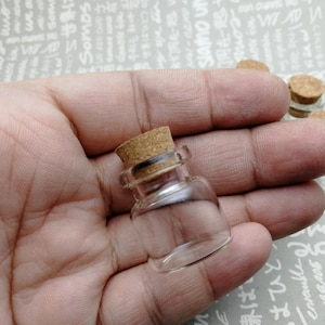 May include: A close-up of a hand holding a small, clear glass bottle with a cork stopper. The bottle is empty and appears to be made of thick glass.