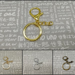 May include: Gold-colored keychain hardware with a lobster clasp, a split ring, and a short chain. The hardware is designed for attaching keys or charms. Additional examples of the same hardware in silver and bronze are shown below the main image.