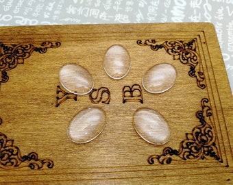Pode incluir: Seis cabochões ovais de vidro transparente sobre uma superfície de madeira com uma borda decorativa. Os cabochões estão dispostos em círculo com as letras "YSB" no centro.