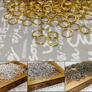 May include: A close-up image of three piles of jump rings. The first pile is gold, the second is silver, and the third is black. The jump rings are all the same size and shape.