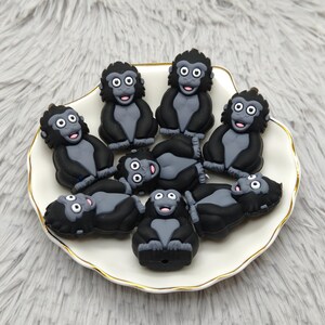 Puede incluir: Una colección de amuletos con forma de mono negro y gris con ojos blancos y bocas rosas, dispuestos sobre un plato blanco festoneado con borde dorado. Los amuletos son de material gomoso.