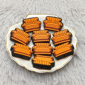 Può includere: Una collezione di ciondoli a forma di divano in miniatura arancione e marrone disposti su un piatto bianco con bordo dorato. I ciondoli sono neri sul fondo e hanno un design dettagliato. Il piatto poggia su una superficie grigia e soffice.