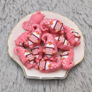 May include: A collection of pink gnome figurines with white polka dots and orange noses, each holding a sign that reads "BEST MOM EVER." The gnomes are arranged in a white and gold-rimmed seashell-shaped dish.