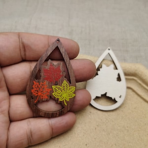 May include: A pair of wooden earrings shaped like teardrops with a cutout design of leaves. One earring is painted with red, orange, and yellow leaves, while the other earring is unpainted.