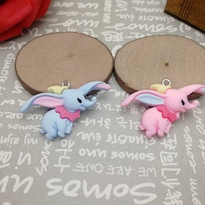 Pode incluir: Dois pingentes de elefante Dumbo, um azul claro e outro rosa, com orelhas grandes e gola com babados. Cada pingente tem um pequeno laço de metal para prender a joias ou artesanato. Os pingentes têm aproximadamente 5 cm de altura.
