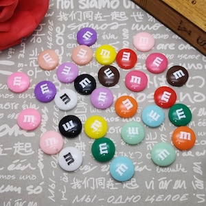 Può includere: Assortimento di decorazioni rotonde piatte in resina colorate con la lettera "M" in bianco. Le decorazioni hanno un diametro di circa 1 cm.