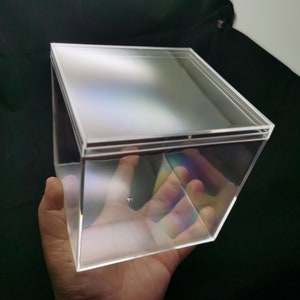 May include: A clear plastic cube-shaped box with a lid. The box is empty and has a slight iridescent sheen.