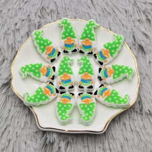 May include: A collection of 12 green and white gnome-shaped resin charms with orange and blue accents. The charms are arranged in a circle on a white plate with a gold rim.