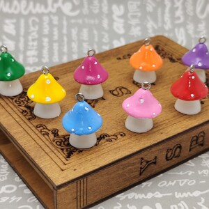 May include: A collection of nine colourful mushroom charms with white polka dots. The charms are displayed on a wooden box with the word "Y" engraved on the front.