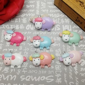 May include: A collection of colorful sheep-shaped charms in various pastel shades, including pink, blue, green, and purple. Each charm has a small silver loop for attaching to jewelry or other crafts. The sheep have smiling faces.