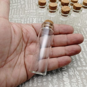 May include: A close-up of a clear glass bottle with a cork stopper. The bottle is held in a hand, and the background is a white surface with black text.