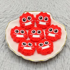 Puede incluir: Siete figuras rojas, con forma de dibujos animados, con mascarillas blancas y ojos negros, dispuestas sobre un plato blanco con volantes y borde dorado. Las figuras tienen forma circular y texturizada. El plato está sobre una superficie gris y difusa.