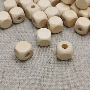 May include: A group of natural wood square beads with a hole in the center. The beads are scattered on a light brown fabric surface.