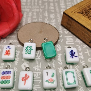 May include: A collection of ten small, white ceramic mahjong tiles with green and red accents. The tiles feature Chinese characters and symbols, including the numbers 1, 8, and 9, as well as the characters for 'wind' and 'middle'.