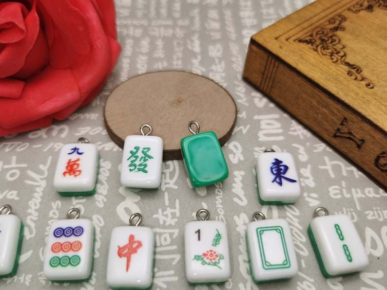 May include: A collection of ten small, white ceramic mahjong tiles with green and red accents. The tiles feature Chinese characters and symbols, including the numbers 1, 8, and 9, as well as the characters for 'wind' and 'middle'.