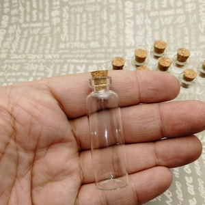 May include: A clear glass bottle with a cork stopper, held in a hand. Several similar bottles are visible in the background. The bottles are small, cylindrical, and appear to be empty.
