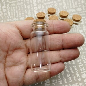 Puede incluir: Una pequeña botella de vidrio transparente con un tapón de corcho. La botella es de forma cilíndrica.