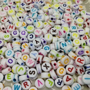 以下が含まれることがあります： カラフルな文字とストライプが付いた白いプラスチック製のアルファベットビーズの詰め合わせです。ビーズは、クラフトプロジェクト、ジュエリー作り、教育活動に最適です。