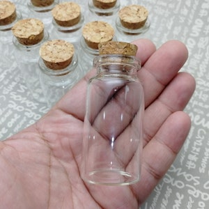 May include: A clear glass bottle with a cork stopper. The bottle is empty and has a round shape. The bottle is being held in a hand.