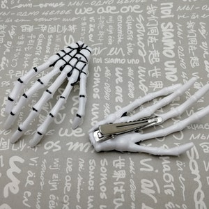 Puede incluir: Dos pinzas para el pelo de plástico blanco en forma de mano de esqueleto. Las pinzas están detalladas con líneas negras para parecerse a los huesos. Las pinces están sobre un fondo gris con texto blanco.