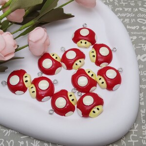 May include: A selection of red mushroom charms with white spots and yellow detailing. Each charm has a silver loop for attaching to jewellery or crafts. The charms are displayed on a white, curved surface.
