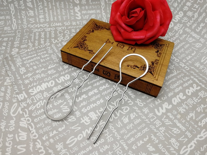 Puede incluir: Tres horquillas de pelo plateadas, dos con un dise&ntilde;o ondulado y una con un bucle circular en el extremo. Las horquillas est&aacute;n sobre una superficie de madera con una rosa roja en el fondo.