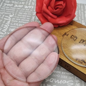 May include: A clear, round magnifying glass is held in a hand. A wooden box with a decorative glass oval and a red rose are in the background. The glass oval has the letters "SB" and floral designs.